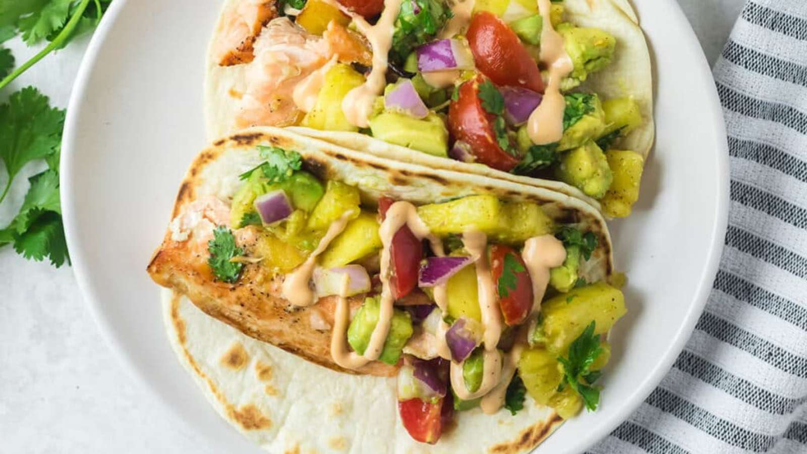 Two soft tacos filled with grilled chicken, diced pineapple, tomato, red onion, avocado, cilantro, and drizzled with a creamy sauce, served on a white plate.