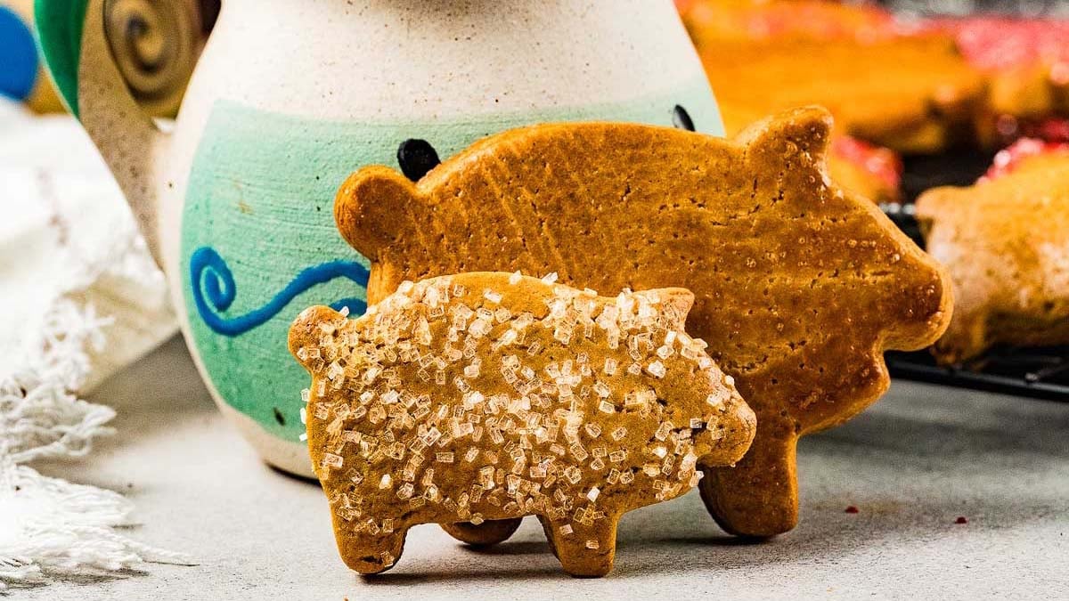 Two pig-shaped cookies, one plain and one topped with coarse sugar, are placed in front of a ceramic mug with blue and green decoration.