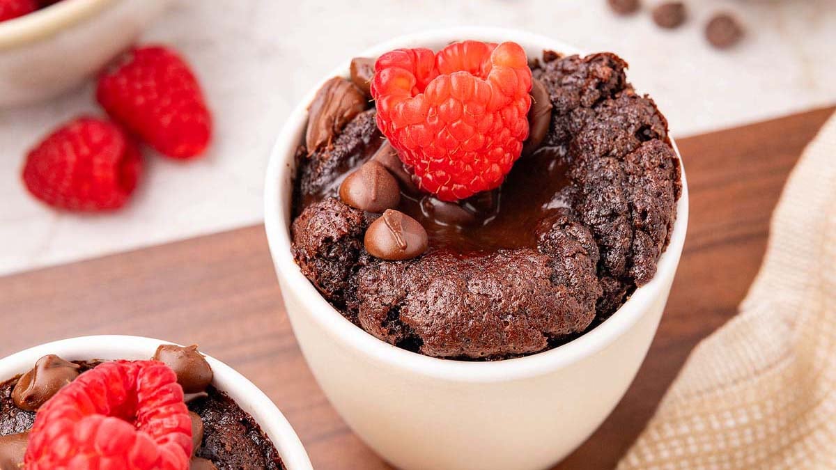 A white ramekin filled with chocolate lava cake, topped with chocolate chips and fresh raspberries, sits on a wooden surface.