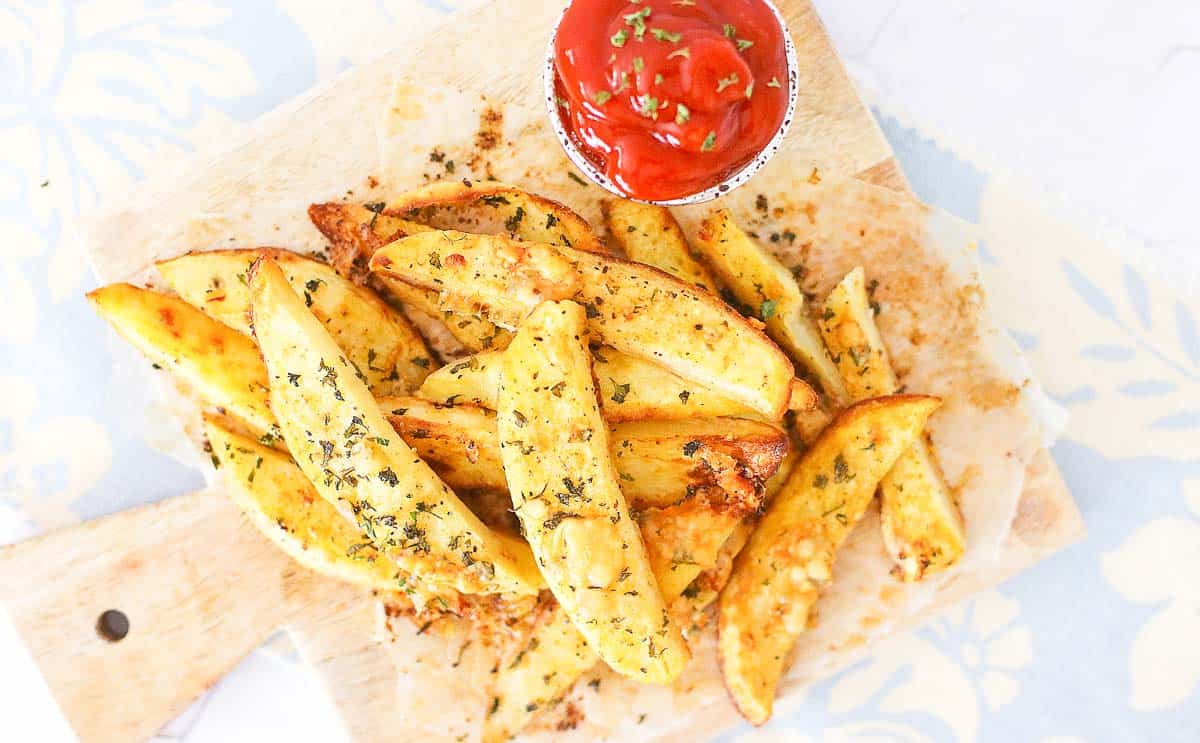 Potato wedges seasoned with herbs on a wooden board, served with a small cup of ketchup garnished with chopped herbs.