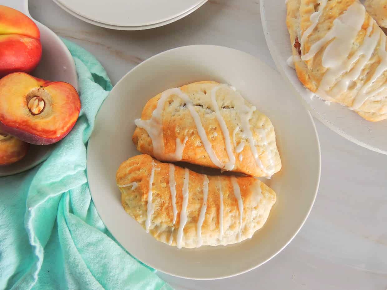 Two glazed turnovers are served on a white plate, with a bowl of fresh peaches and a turquoise cloth nearby on a light-colored surface.