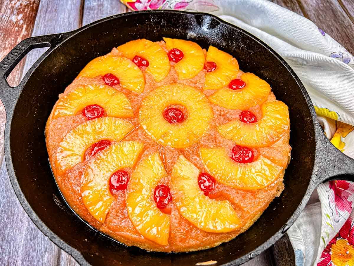 A pineapple upside-down cake topped with pineapple rings and maraschino cherries in a black cast iron skillet on a wooden surface.