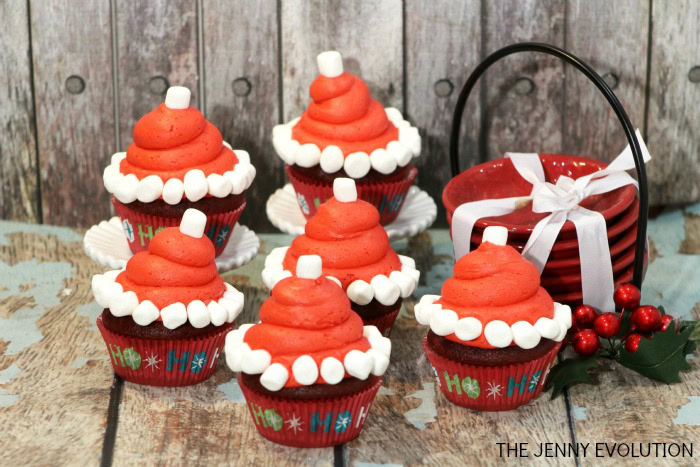 Six cupcakes decorated with red frosting, mini marshmallows, and a single marshmallow on top, resembling Santa hats, are arranged on a wooden surface next to red stacked bowls tied with a white ribbon.