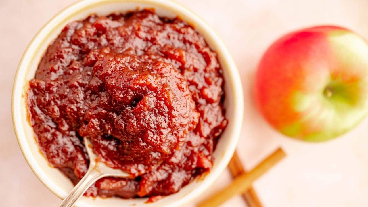 A bowl of thick, dark brown apple butter with a spoon, next to a red and green apple and two cinnamon sticks on a light surface.
