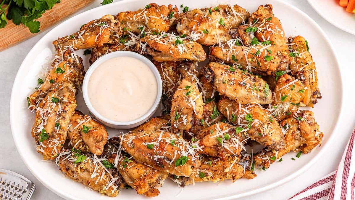 A white platter of baked chicken wings garnished with chopped herbs and grated cheese, arranged around a bowl of creamy dipping sauce.