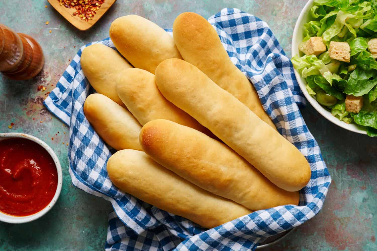 A basket of breadsticks on a blue and white checkered cloth next to a bowl of salad with croutons and a dish of marinara sauce.