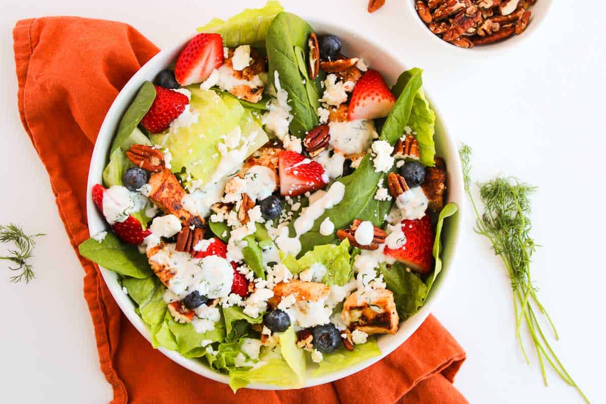 A bowl of salad with grilled chicken, strawberries, blueberries, pecans, lettuce, and crumbled cheese, topped with creamy dressing, on an orange napkin.