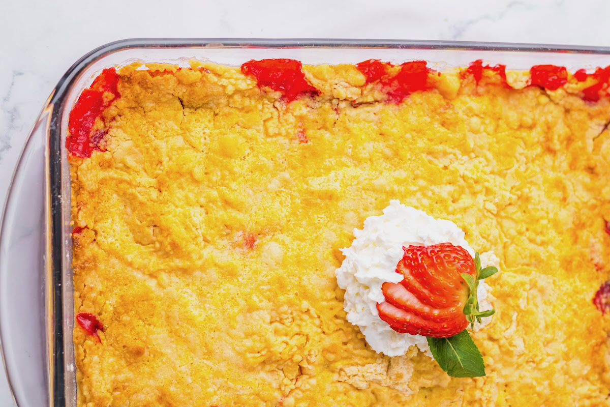 A baked dessert with a golden crust in a glass dish, topped with whipped cream, sliced strawberries, and a mint leaf.