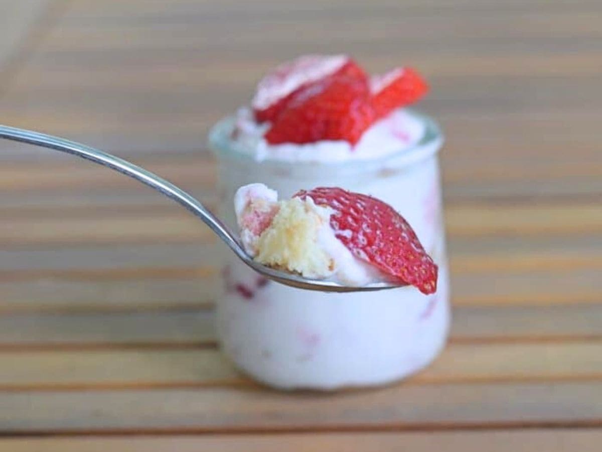 A spoonful of yogurt with strawberries and a bit of cake is held in front of a small glass jar containing the same dessert, on a wooden surface.