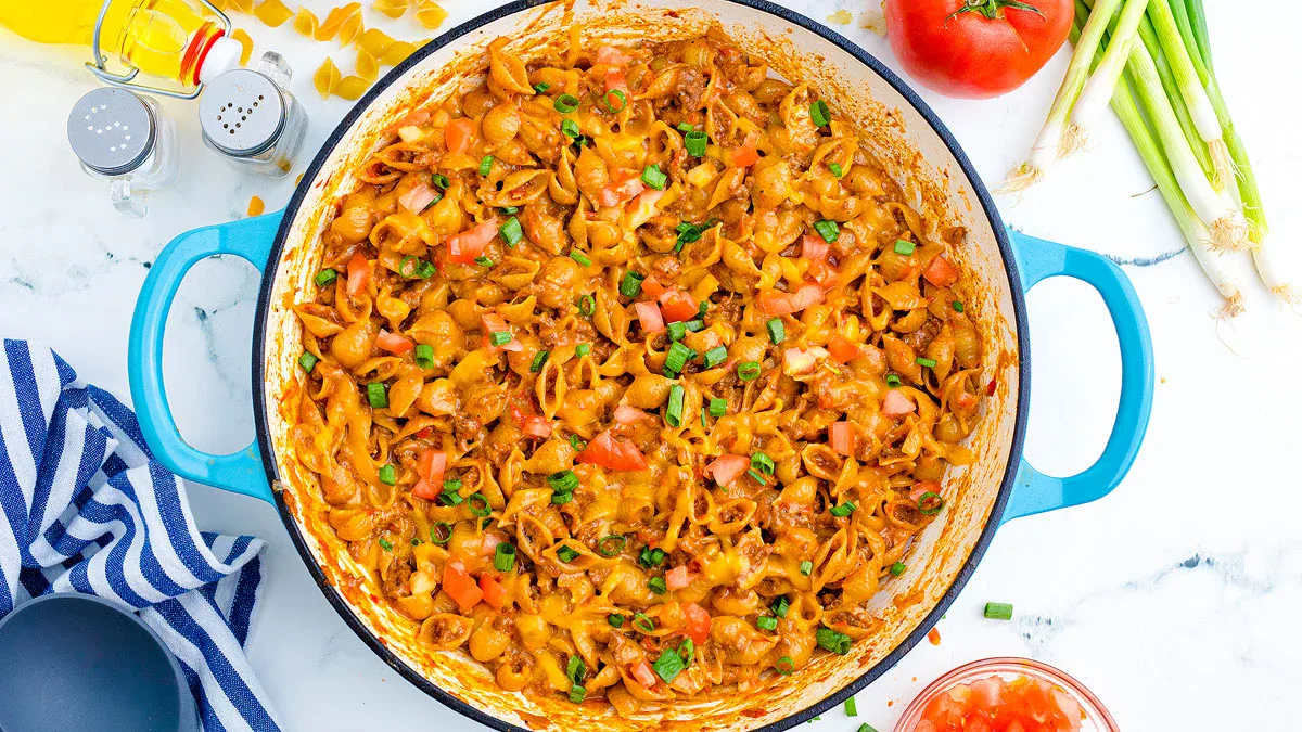 A blue pot filled with cheesy taco pasta topped with chopped tomatoes and green onions, surrounded by fresh ingredients on a white countertop.