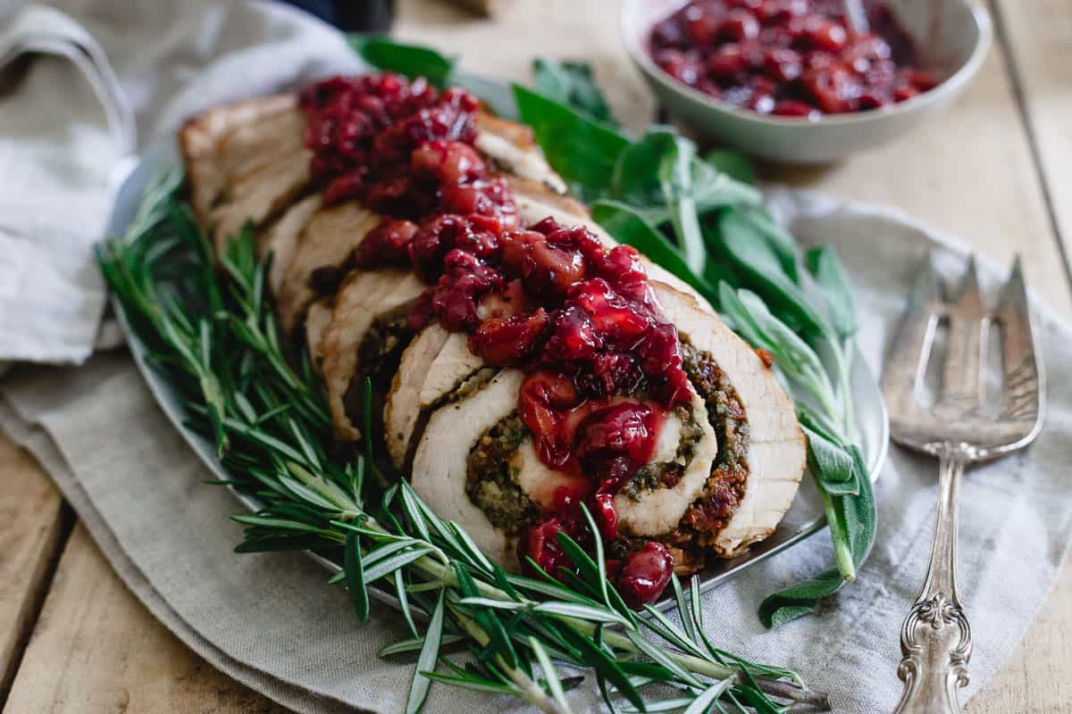 Sliced stuffed pork loin garnished with fresh herbs and topped with cranberry sauce, served on a platter with a bowl of extra sauce beside it.