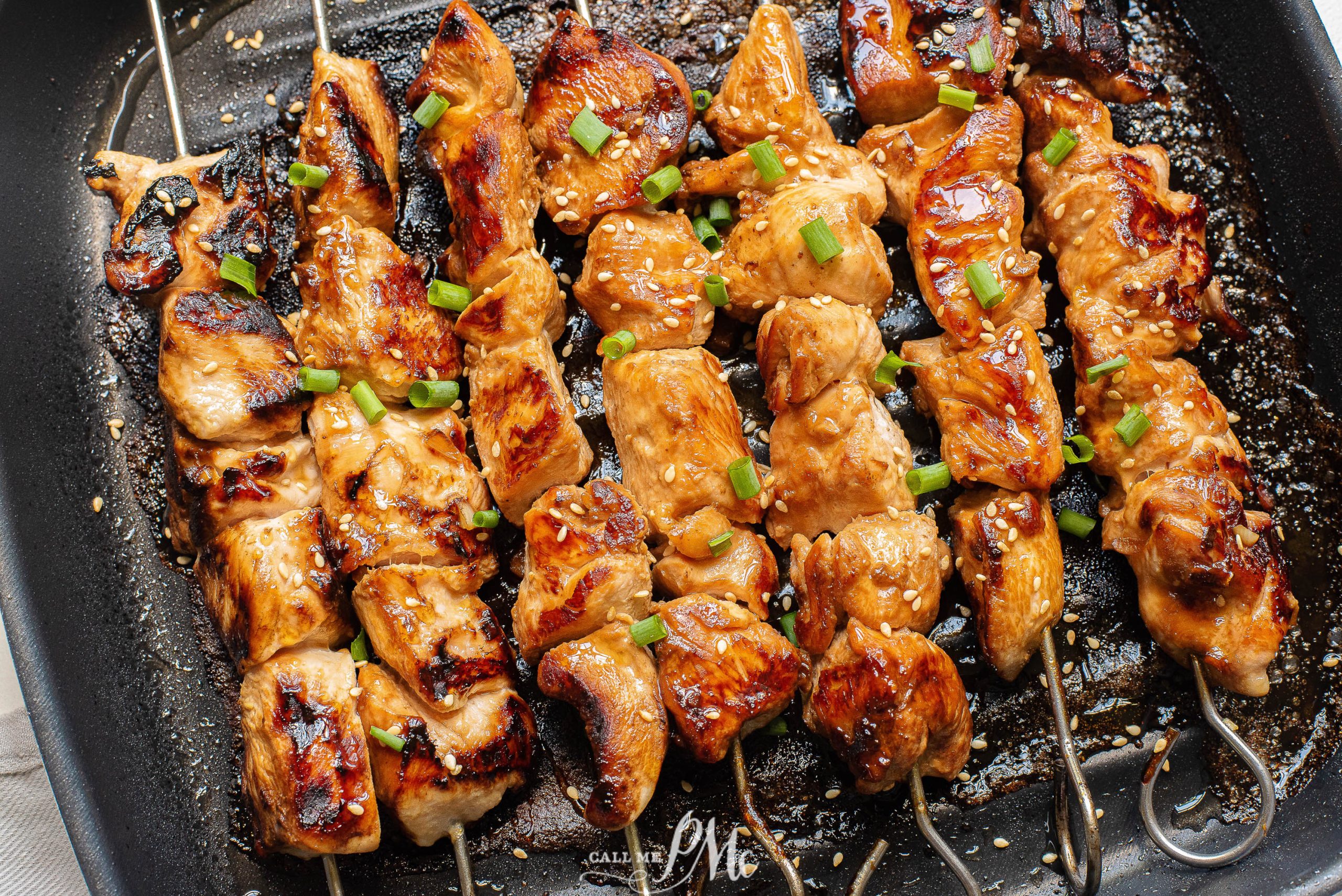 Four grilled chicken skewers topped with chopped green onions and sesame seeds on a grill pan.