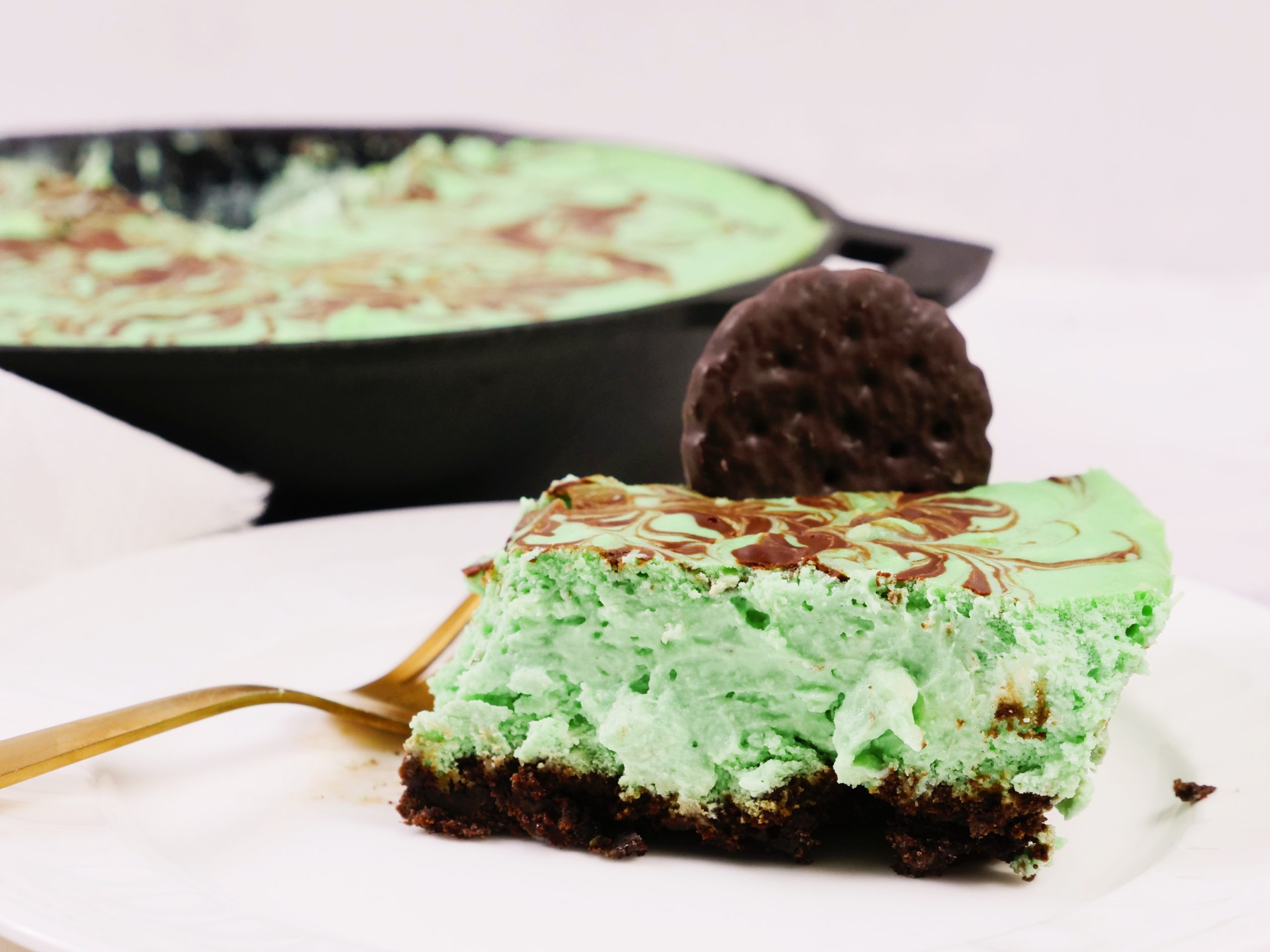 A slice of green mint cheesecake with a chocolate cookie base is served on a white plate with a gold fork; a mint chocolate cookie and the rest of the cheesecake are in the background.