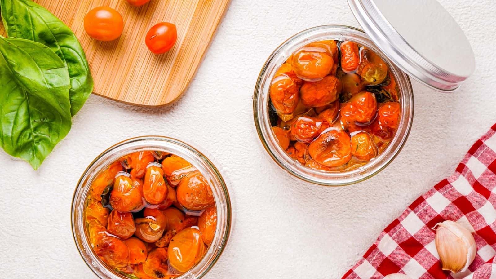 Two glass jars filled with cherry tomatoes in oil are on a white surface, next to fresh basil leaves, a wooden board with cherry tomatoes, a red checked cloth, and a garlic clove.