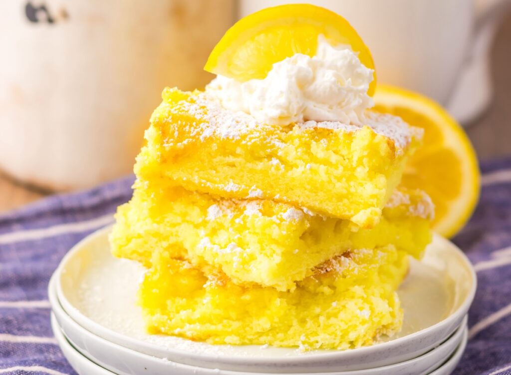 Three stacked lemon bars topped with whipped cream and a lemon slice, dusted with powdered sugar, on a white plate with a striped cloth underneath.