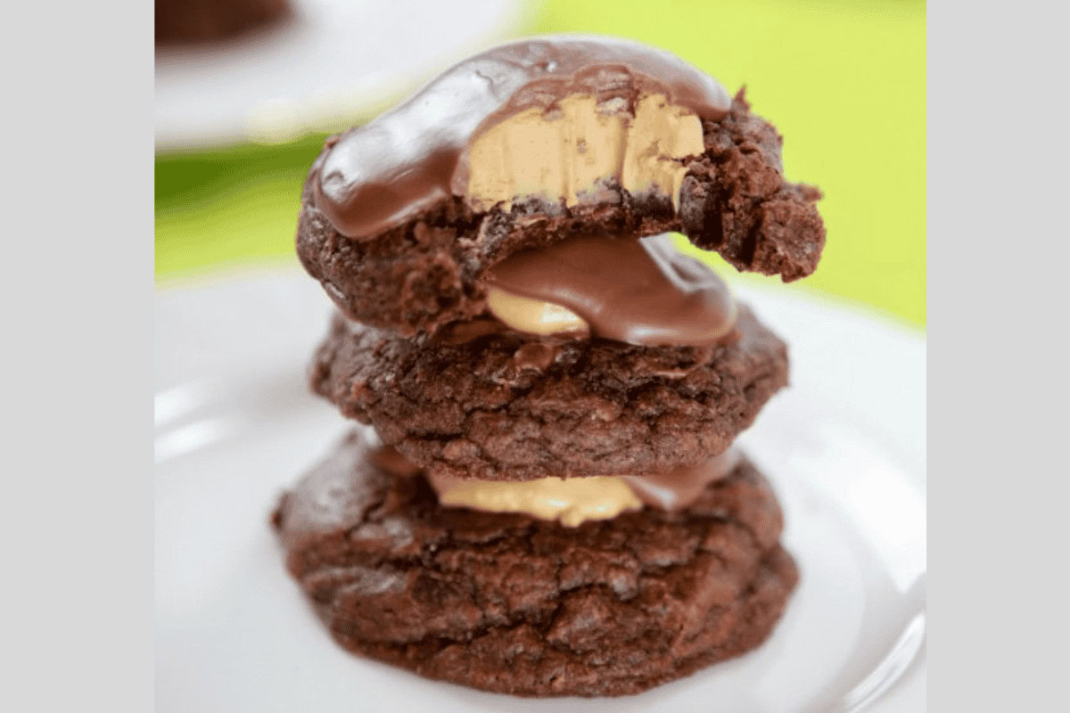 A stack of three chocolate cookies with a creamy filling and chocolate coating, with the top cookie partially bitten, on a white plate.