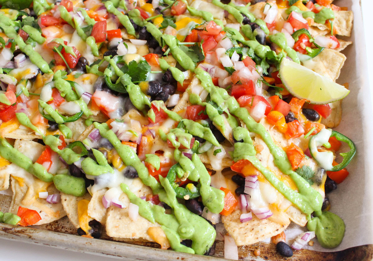 A metal sheet pan topped with cheesy nachos, black beans, bell peppers, and avocado crema.