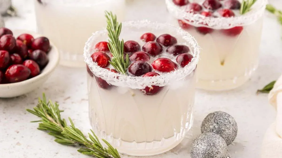 A glass of white cocktail garnished with cranberries and a rosemary sprig, rimmed with sugar, sits on a light surface beside more cranberries, rosemary, and silver ornaments.