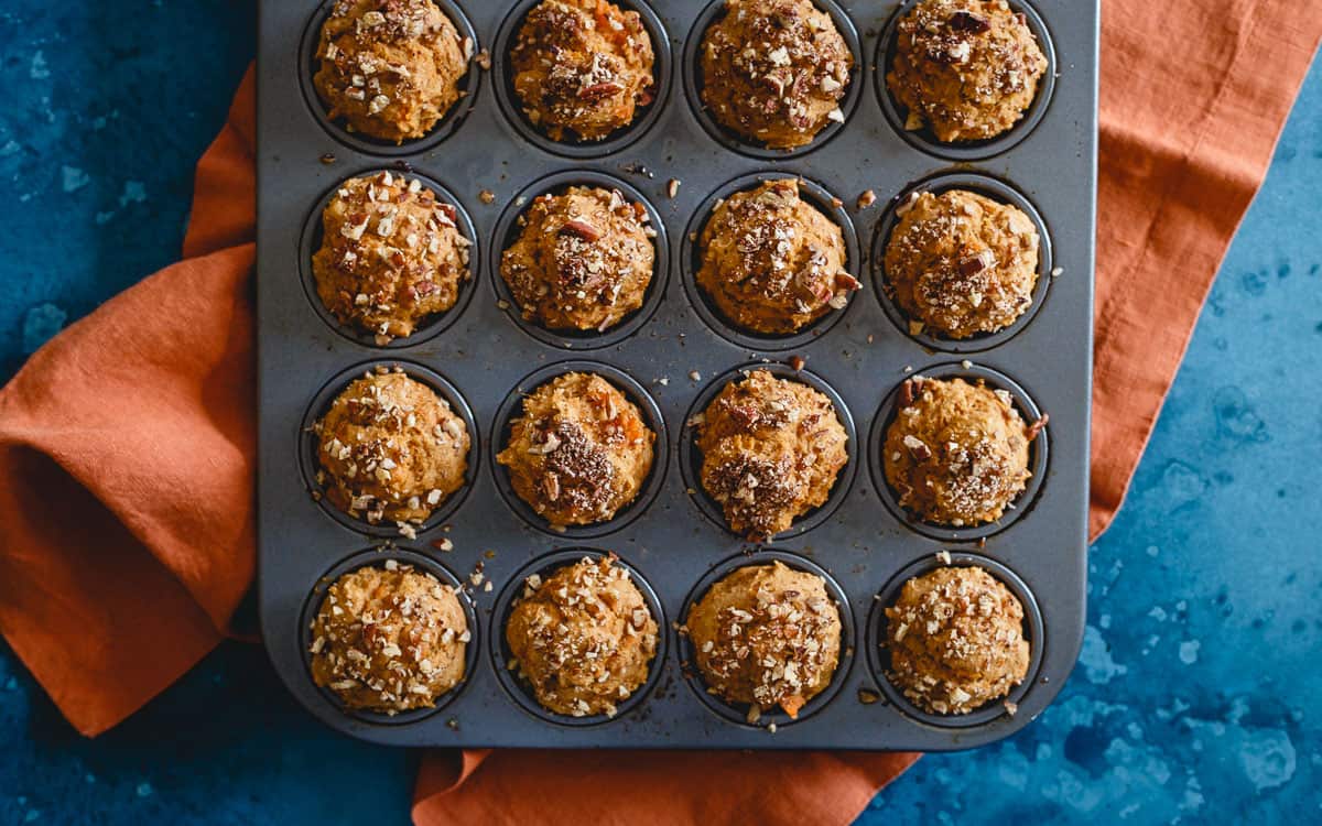 A muffin tin filled with sixteen baked muffins topped with a crumbly streusel, placed on an orange cloth on a blue surface.