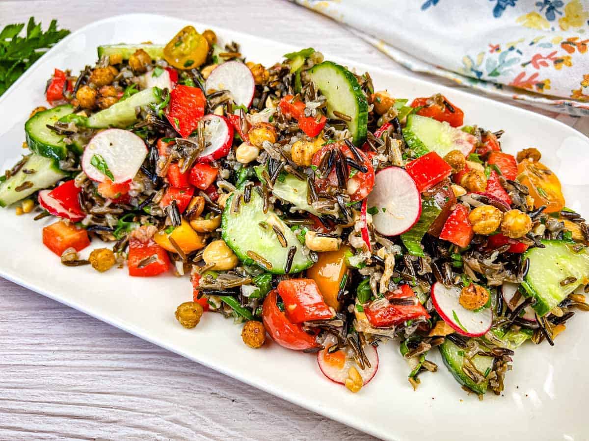 A plate of wild rice salad with chopped cucumbers, radishes, tomatoes, red bell peppers, chickpeas, and fresh herbs on a white rectangular dish.