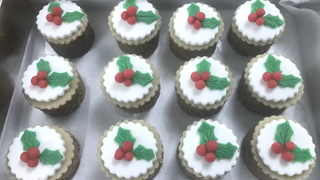 Twelve cupcakes decorated with white icing and green holly leaves with red berries, arranged in rows on a tray.
