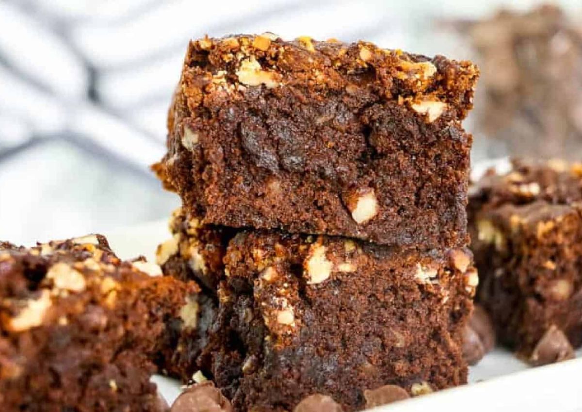 Close-up of chocolate brownies with chopped nuts stacked on a white plate, with more brownies visible in the background.