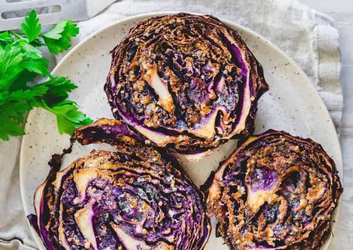 Three roasted cabbage steaks with browned edges are arranged on a white plate, with a sprig of fresh parsley nearby.