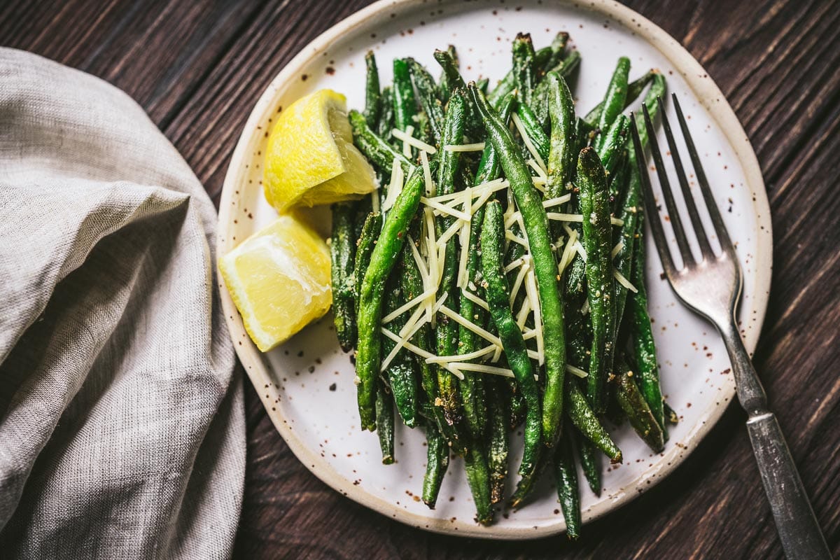 A plate of cooked green beans topped with shredded cheese, served with two lemon wedges and a fork on the side.