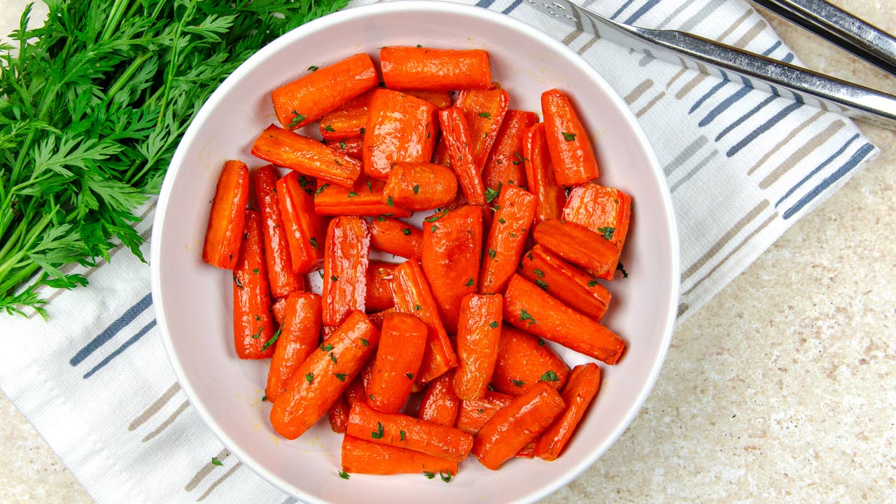 A white bowl filled with roasted carrot pieces garnished with herbs sits on a striped cloth next to a bunch of fresh carrot tops and metal utensils.