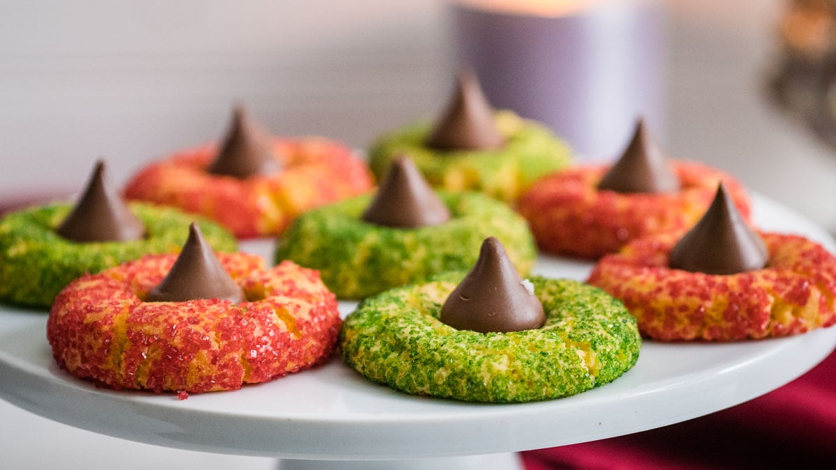 A plate of red and green sugar-coated cookies, each topped with a chocolate kiss, displayed on a white stand.