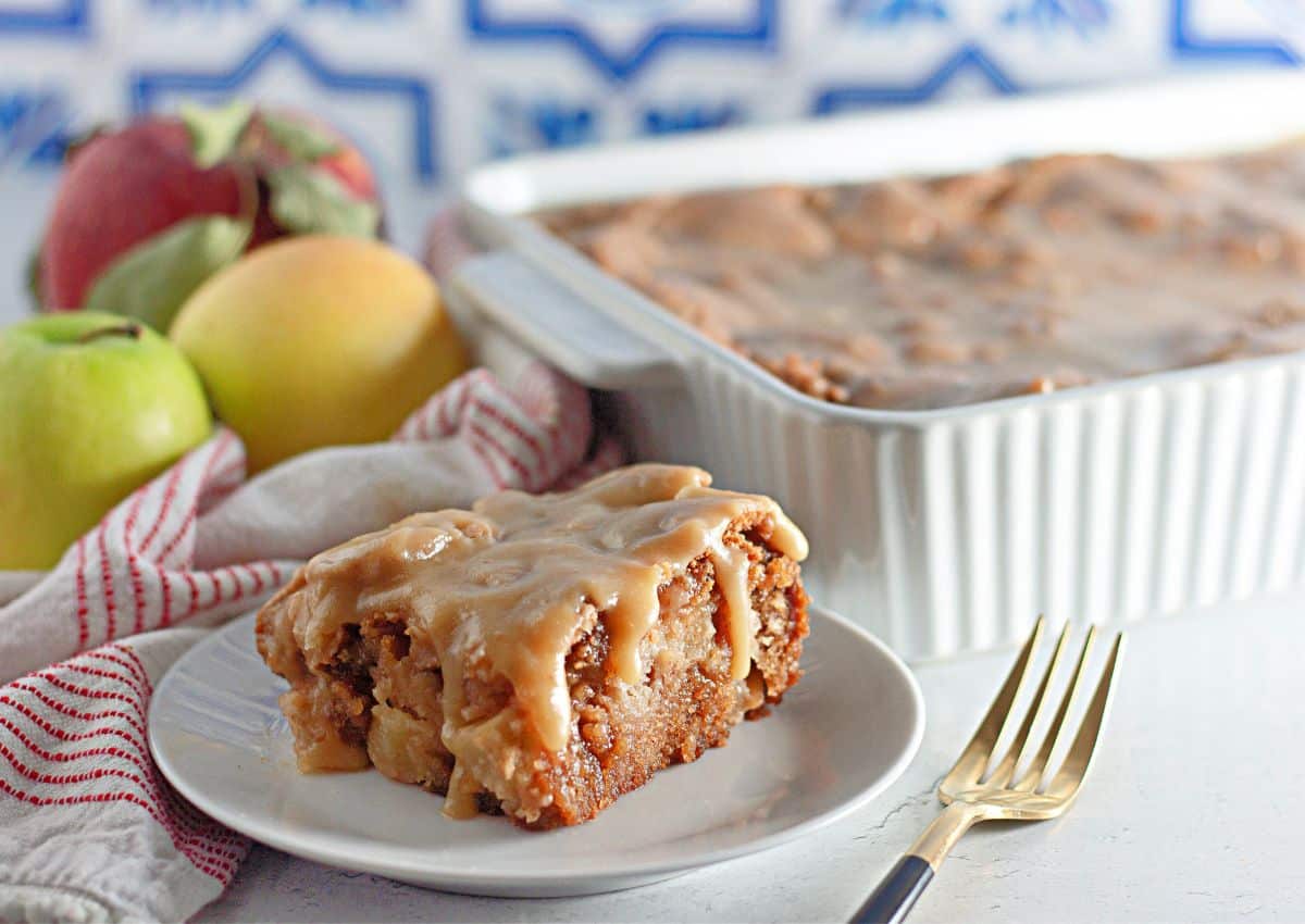 A slice of glazed apple cake sits on a white plate with a fork; a baking dish and whole apples are in the background.
