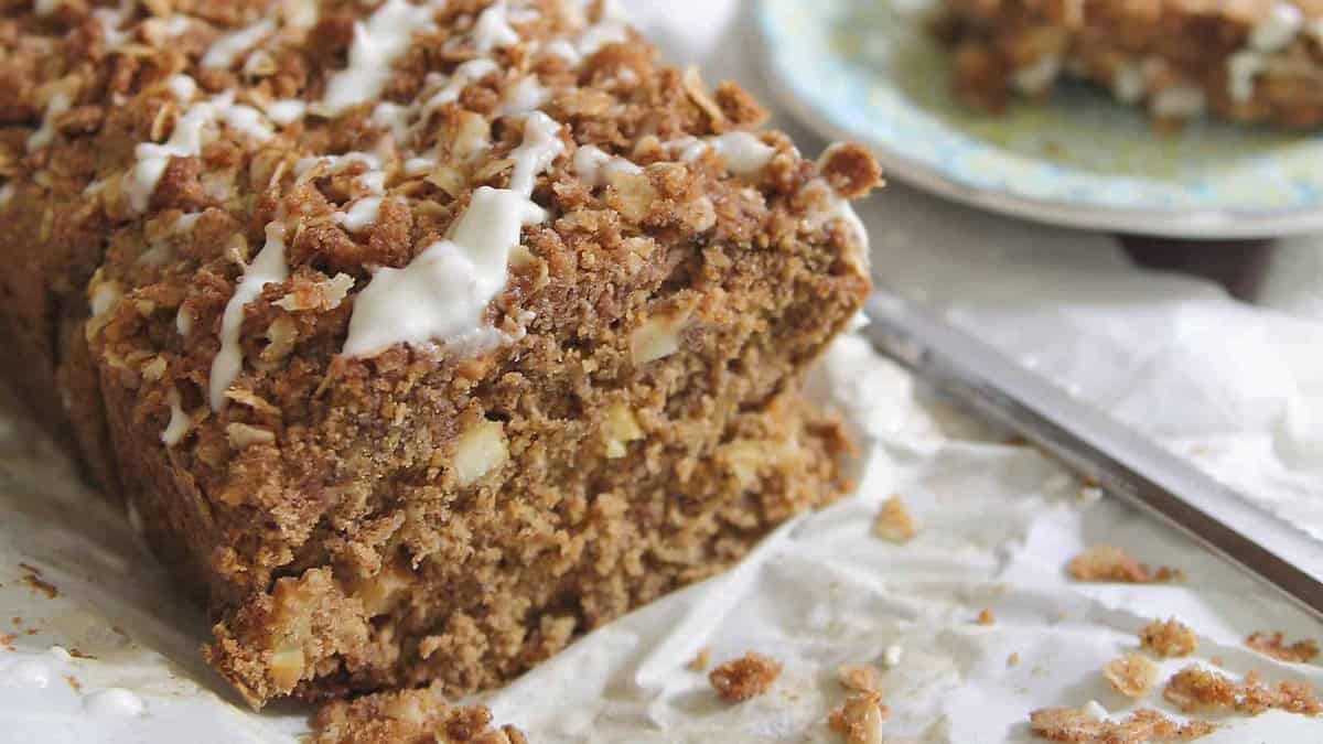 A loaf of crumb-topped apple bread with icing drizzle sits on parchment paper, with a partial slice on a plate in the background.