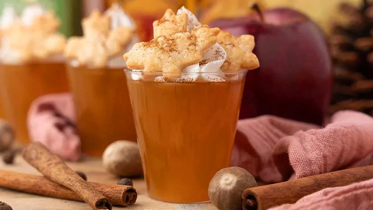 A glass of apple cider garnished with whipped cream, a leaf-shaped pastry, and sprinkled cinnamon, surrounded by cinnamon sticks, a nutmeg seed, and an apple.