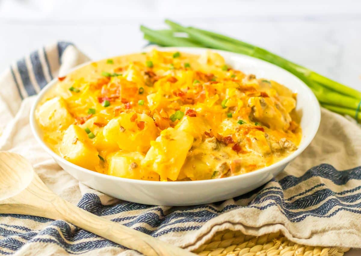 A white bowl filled with cheesy potato casserole, garnished with chopped green onions, sits on a striped cloth next to a wooden spoon and fresh green onions.