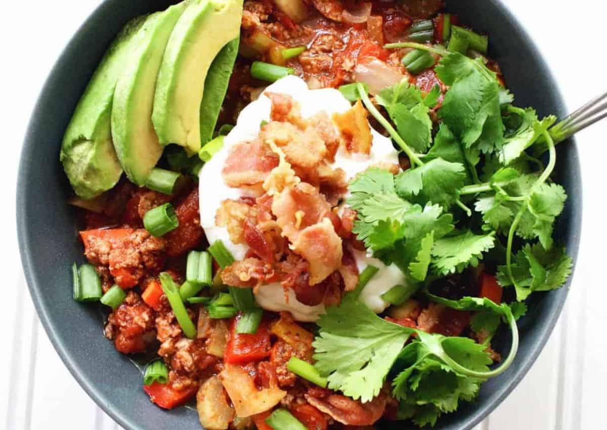 A bowl of chili topped with sliced avocado, sour cream, crumbled bacon, chopped green onions, and fresh cilantro.
