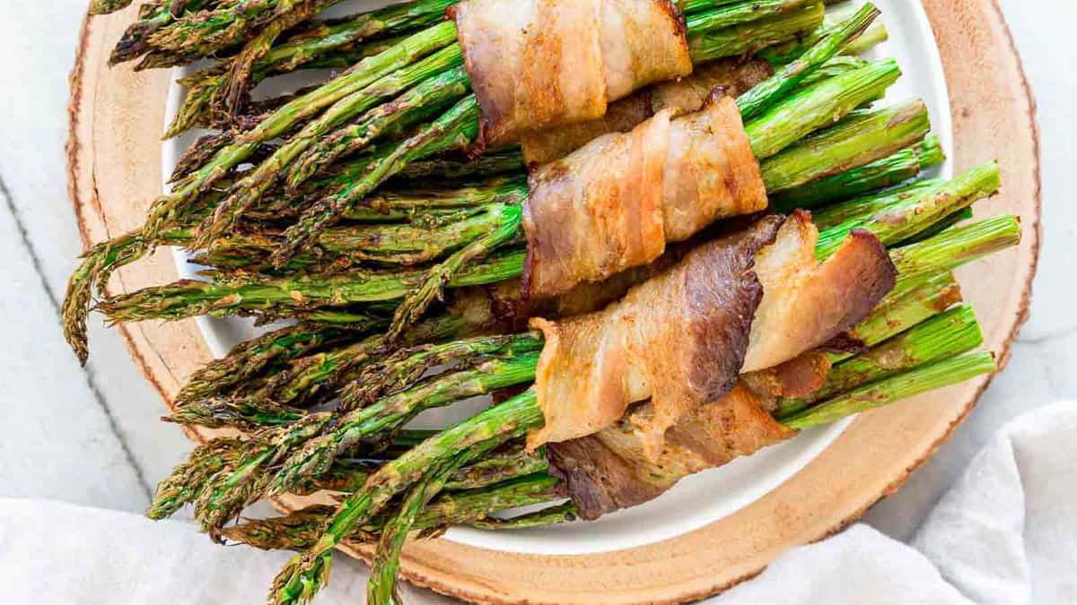 A plate of asparagus spears wrapped in crispy bacon, served on a white dish with a wooden base.