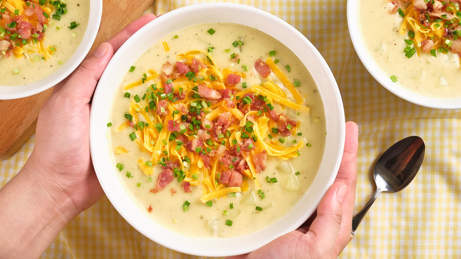 A person holds a bowl of creamy potato soup topped with shredded cheese, bacon bits, and chopped chives. A spoon and two more bowls of soup are nearby on a checkered tablecloth.