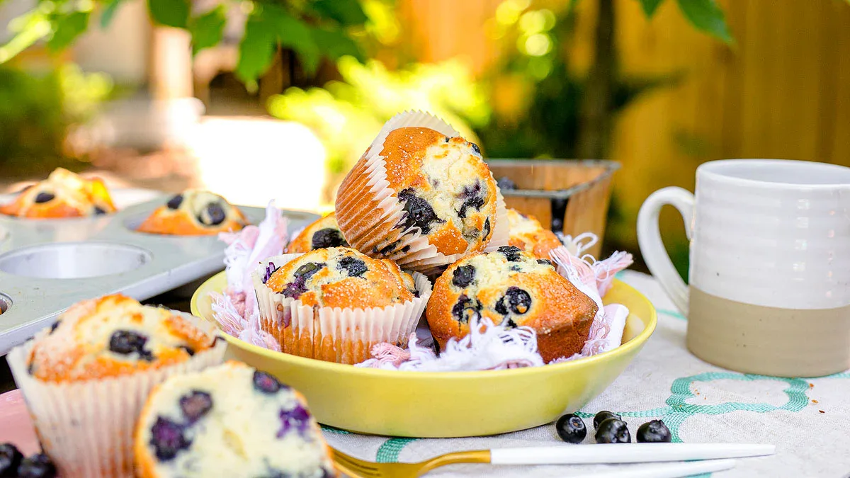 A plate of blueberry muffins sits on an outdoor table with a white mug, napkin, and fork, with more muffins and greenery in the background.