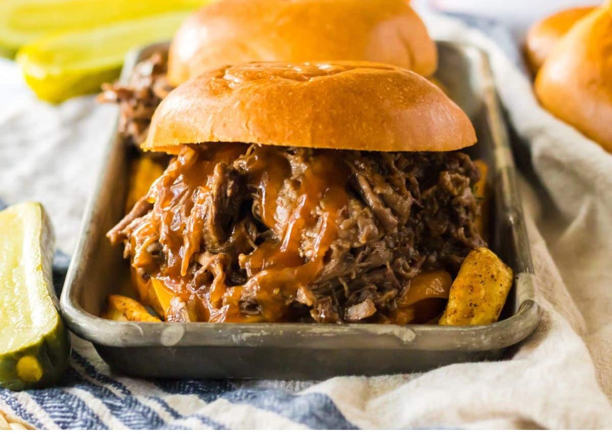 A shredded beef sandwich with barbecue sauce on a bun, served on a metal tray with seasoned potato wedges and pickle slices.