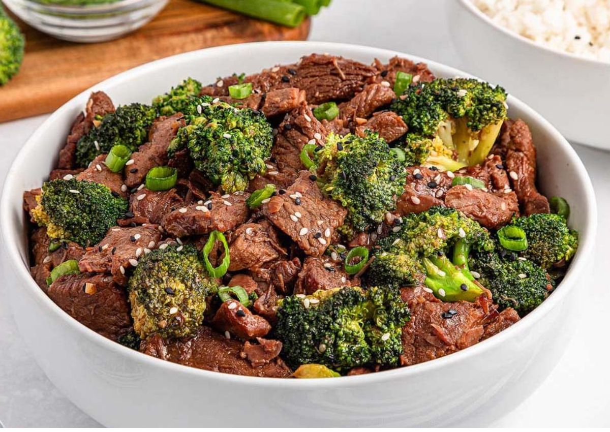 A white bowl filled with beef and broccoli stir-fry, topped with sesame seeds and sliced green onions.