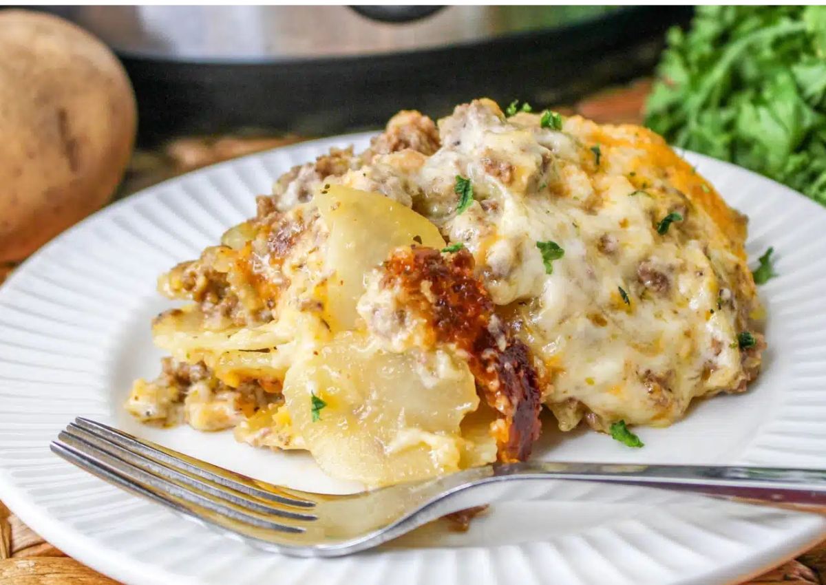 A serving of cheesy ground beef and potato casserole on a white plate with a fork, garnished with chopped herbs.