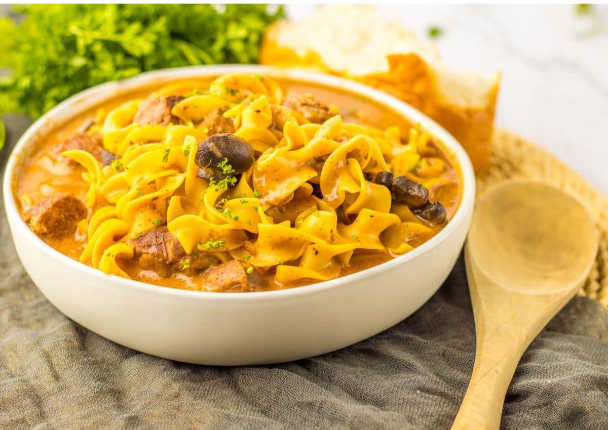 A bowl of creamy beef stroganoff with egg noodles, mushrooms, and parsley garnish, next to a wooden spoon and a piece of bread.