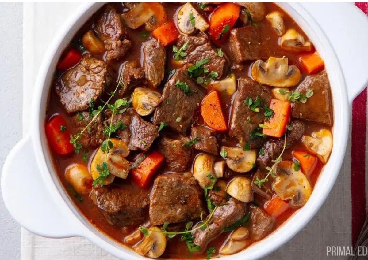 A white bowl filled with beef stew containing chunks of beef, sliced mushrooms, carrots, and garnished with fresh herbs in a rich brown broth.