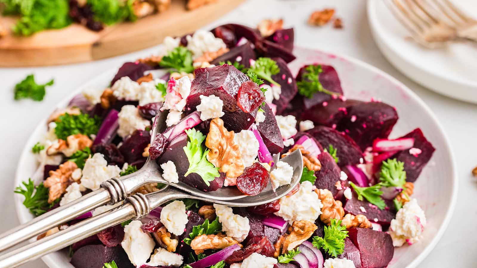 A bowl of beet salad with feta cheese, walnuts, red onions, parsley, and dried cranberries, with a serving spoon lifting a portion.