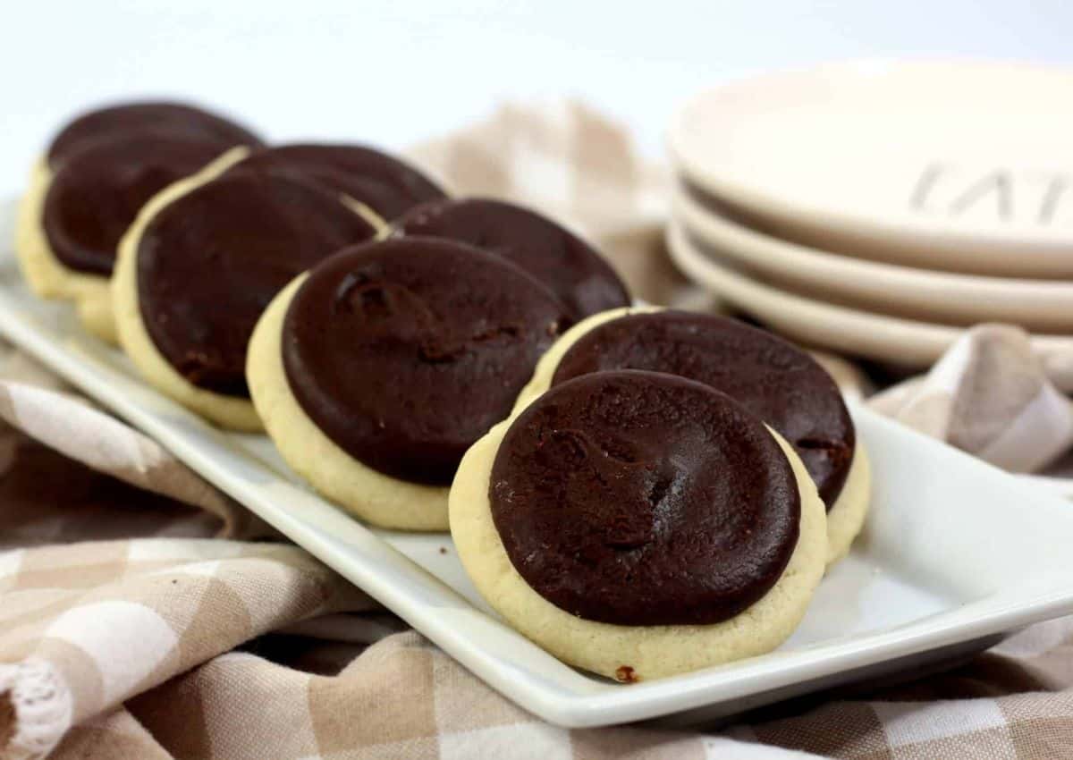 A rectangular white plate holding a row of cookies with chocolate frosting on a beige checkered cloth next to a stack of plates.