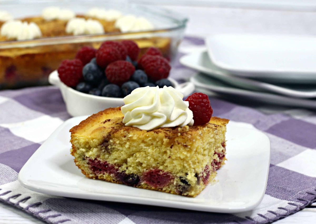 A slice of berry cake with a dollop of whipped cream and a raspberry on top, served on a white plate, with a bowl of mixed berries in the background.