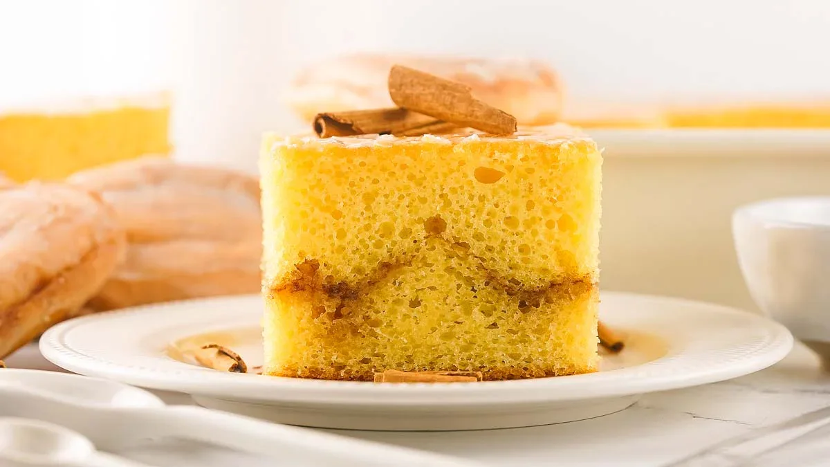 A slice of yellow sponge cake with a cinnamon swirl, topped with cinnamon sticks, is served on a white plate.