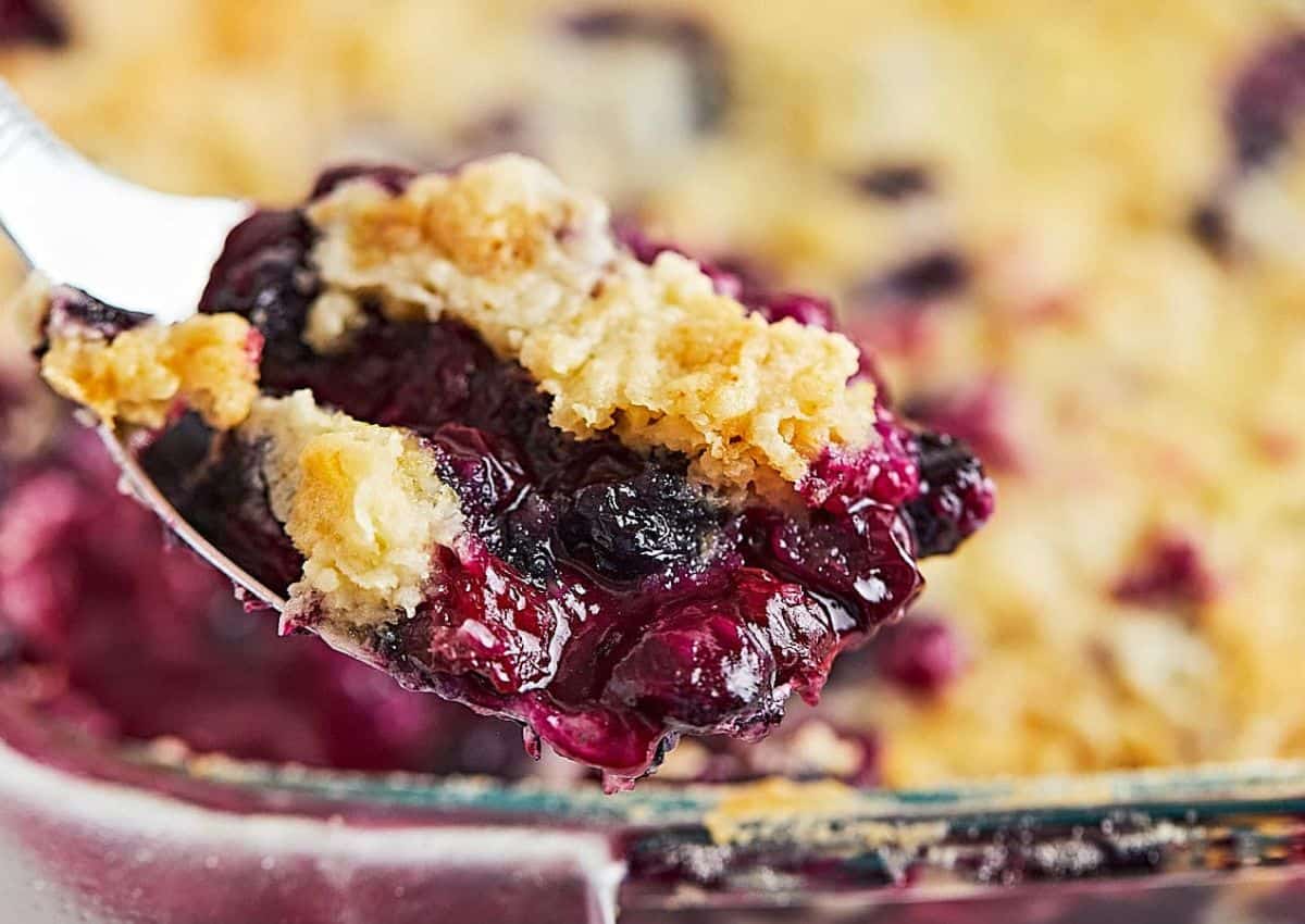 A close-up of a spoonful of blueberry crumble with a golden, crumbly topping and juicy berries, held above a baking dish.