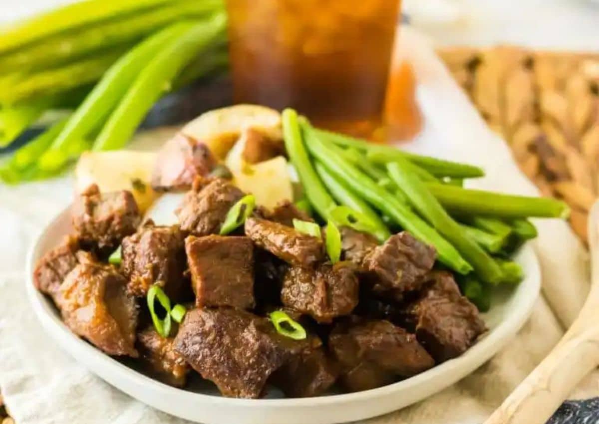 A plate of cooked beef cubes garnished with green onions, served with green beans and roasted potatoes, with a drink in the background.