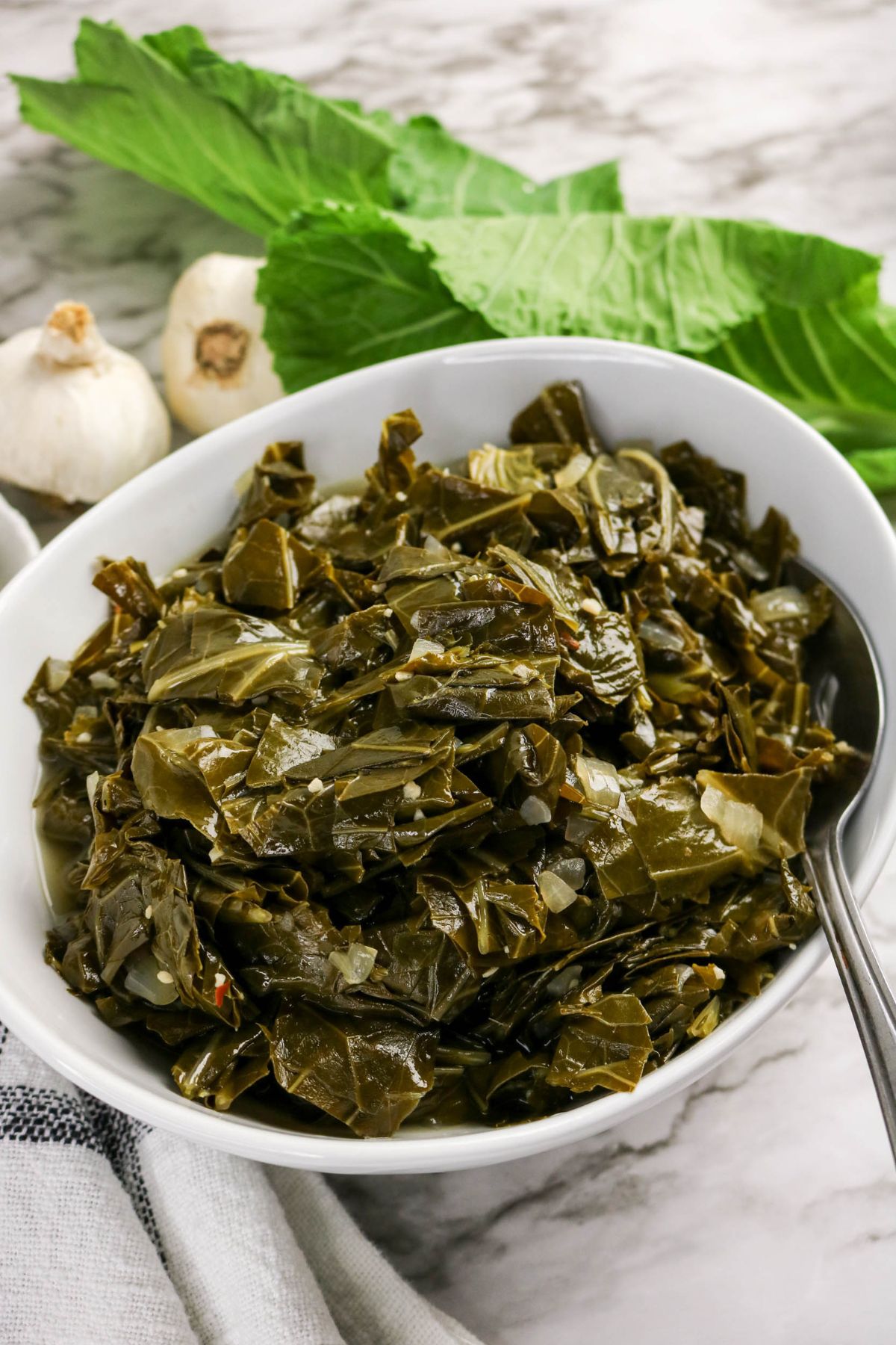 A bowl of cooked collard greens with a spoon, placed on a marble surface next to raw collard leaves and garlic bulbs.